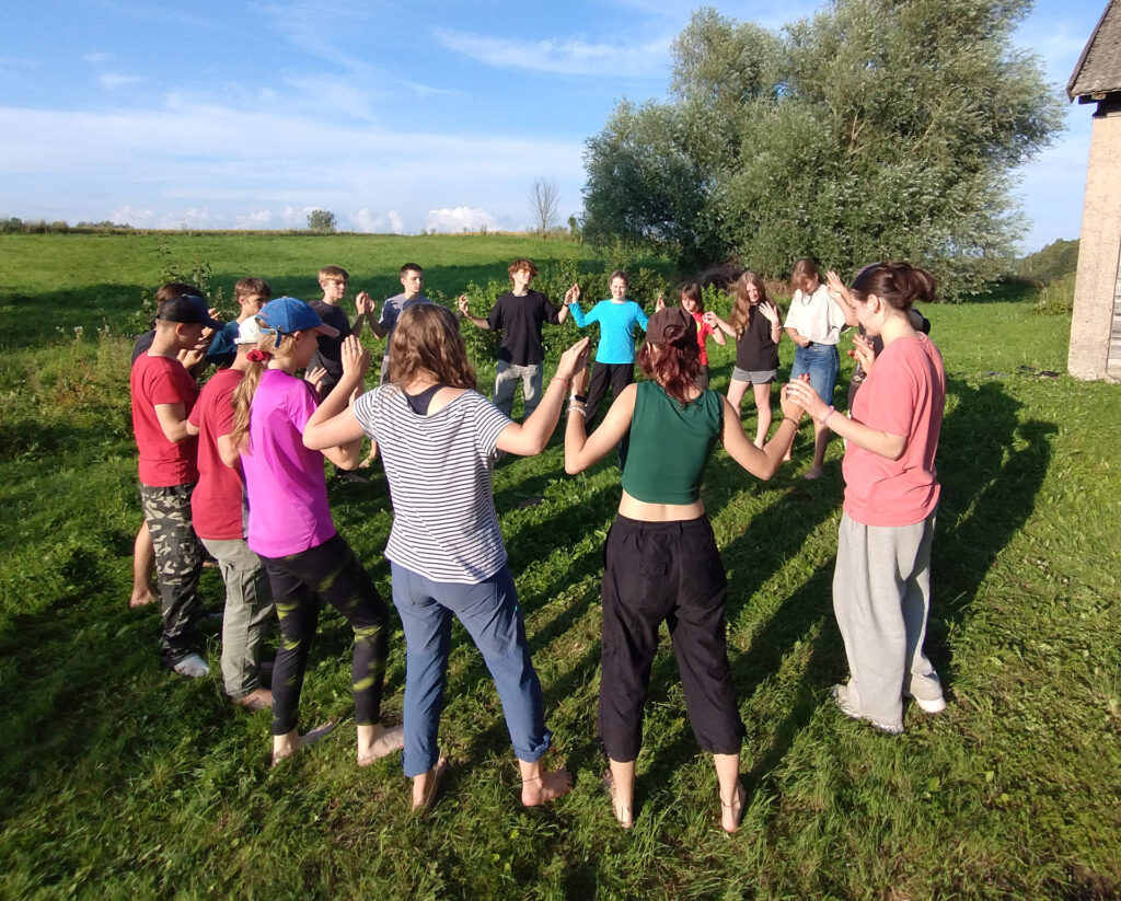 Na łące duża grupa dzieci z wychowawcami stoi w kółku trzymając się za ręce.