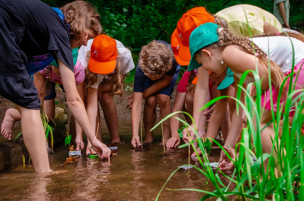 Grupka dzieci stojąc w płytkiej wodzie z zainteresowaniem ogląda coś w jeziorze.